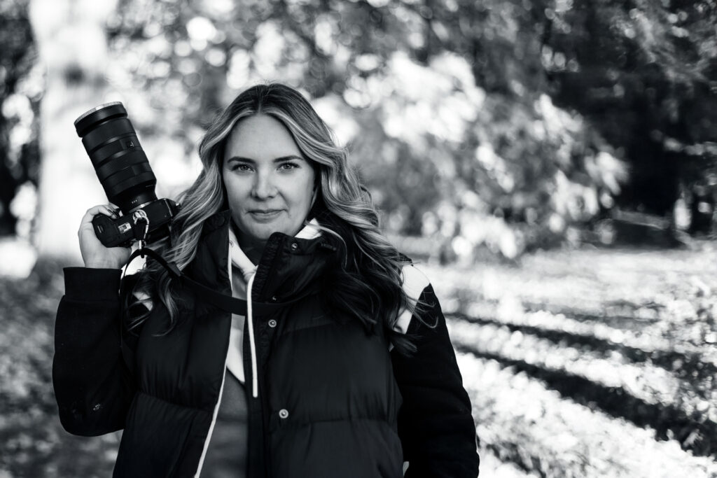 Alicia Riddle confidently holding her camera in a serene black-and-white outdoor forest setting.