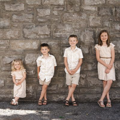 Bohemier siblings posing in light outfits before a rustic stone wall family portrait.