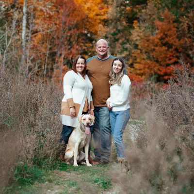 Smiling family with dog in colorful autumn park surrounded by golden and red leaves.