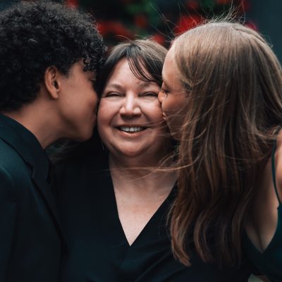 Mother smiling as two children kiss her cheeks during an outdoor family celebration.